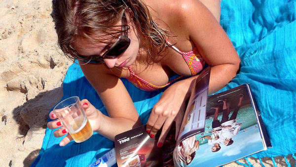 Mulher folheando revista na praia segurando copo de cerveja